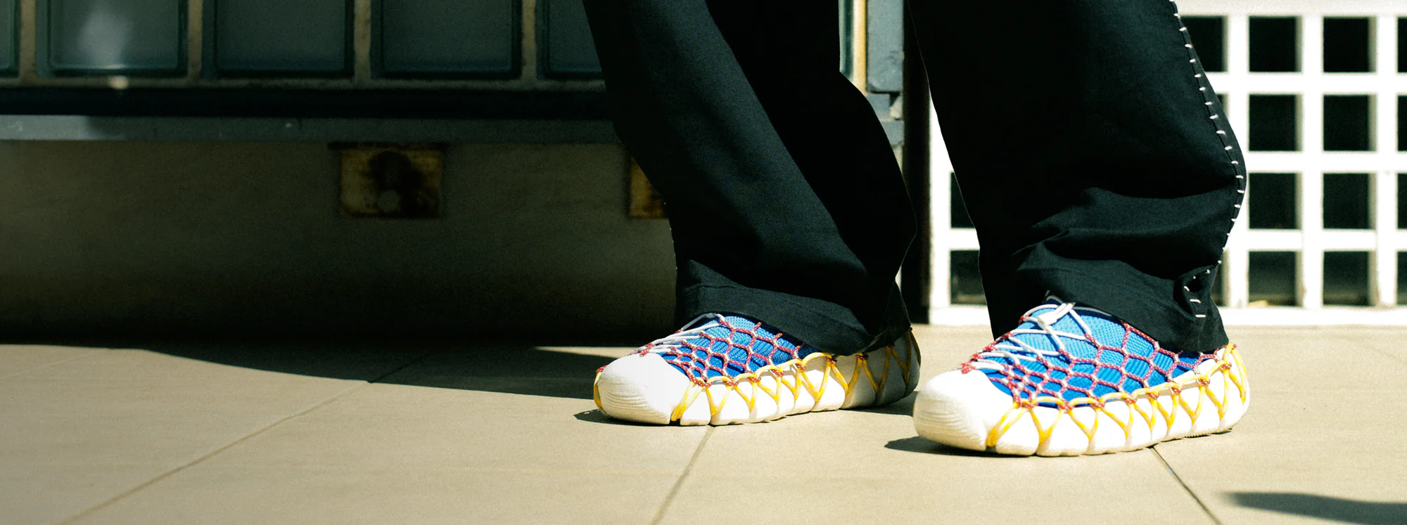 Man in stylish black pants wearing the "Space Available" collaboration Uneek 360 Sneakers in blue and white, detailed with red and yellow lacing.